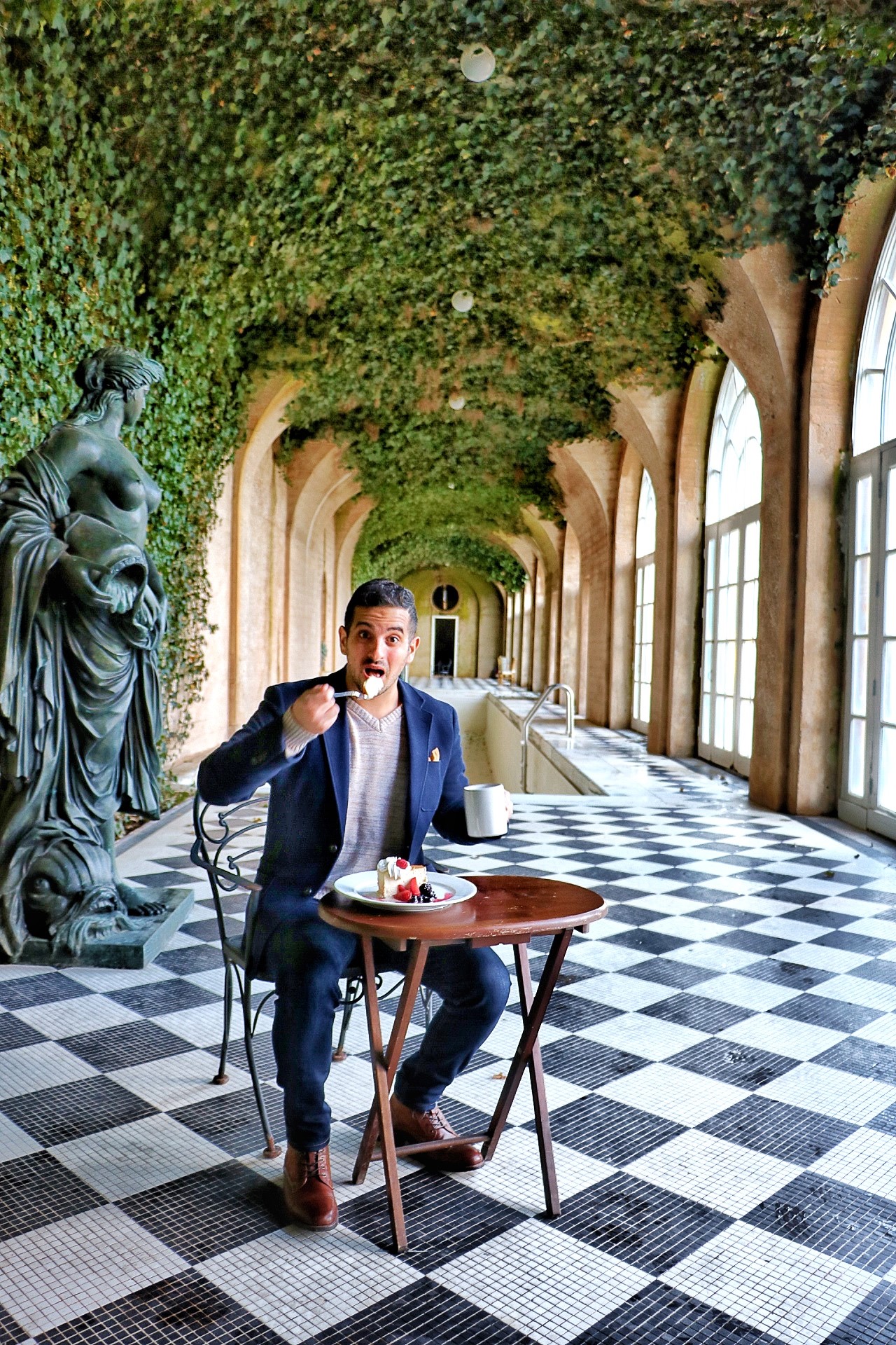 Saint Leo alum Salvatore DiBenedetto, a.k.a. the Grubfather, sitting in a European-inspired, vine-covered, indoor sun room eating dessert and holding a coffee cup